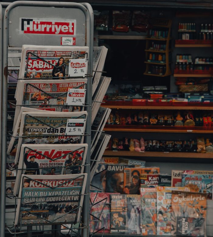 hero-img-01 Metal newspaper stand displaying various Turkish publications at a storefront.