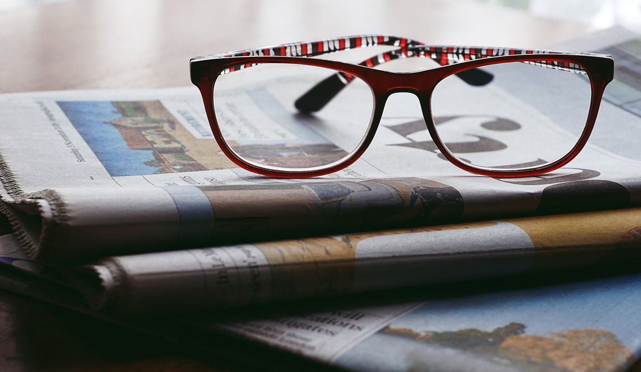 hero-img-02 Close-up of reading glasses resting on a stack of newspapers, symbolizing knowledge and study.